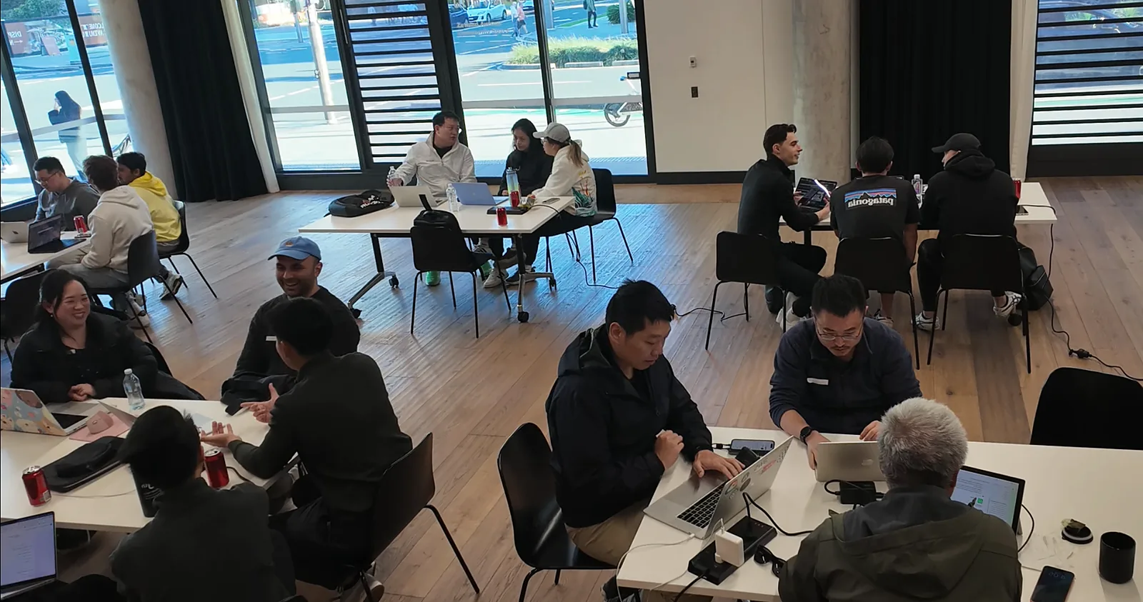 Builders at tables around the room, working on laptops in small groups, with the street visible through floor-to-ceiling windows.