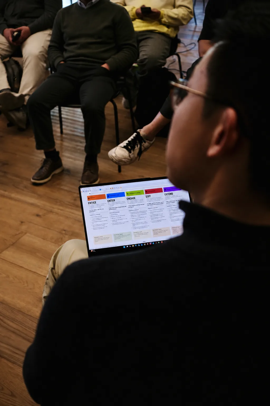 A builder sits with a laptop showing the clinic journey mapped across five colour-coded stages, with the seated room out of focus beyond.
